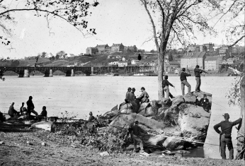 Small group of Civil War soldiers stand near large rocks and look out over Potomac River with Georgetown in the background.