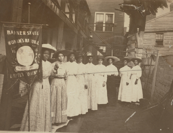 Nine African-American women posed, standing, full length, with Nannie Burroughs holding banner reading, "Banner State Woman's National Baptist Convention"