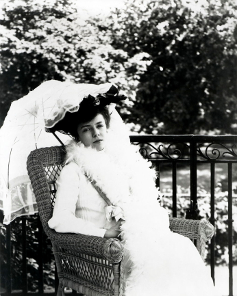 Black and white photo of young woman in white dress and white parasol sitting in chair.