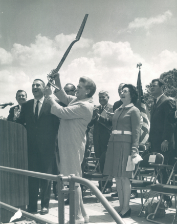 Woman stands on stage and holds shovel above her head as crowd behind her laughs.