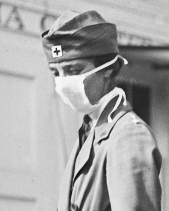 This is a black and white photograph of a woman wearing a mask, she wears a hat with the familiar plus sign associated with the Red Cross. Despite the mask covering most of her face, she wears a serious expression. 