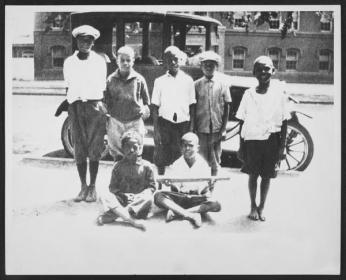 A group of adolescent boys gathered around an old fashioned car in Southwest. Most of the seven boys appear to be African American, but at least one is white. The picture is from 1924. 