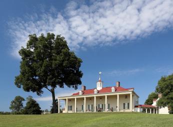 A picture of Mount Vernon from the perspective of the Potomac. A green lawn is seen in the foreground. The mansion is in the background with the Piazza in full view. 
