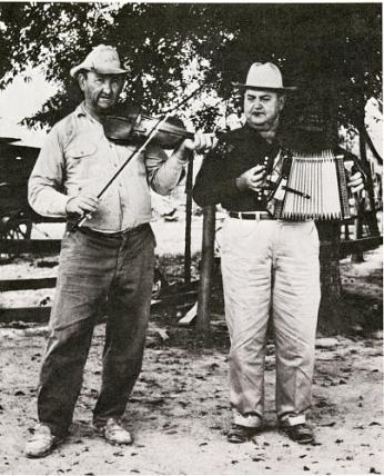 Two men standing in the foreground. The left person is playing a violin while the right person is playing an accordion. 