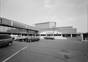 Image of a indoor shopping mall built during urban renewal in Southwest. Early 1970 cars are parked in the foreground while the  shopping complex is in the background making an L shape. 