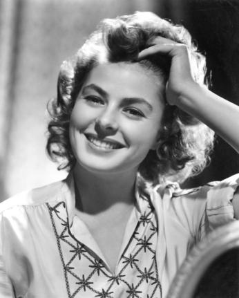 A black and white photograph of Ingrid Bergman. She wears a smile. Her hair is done up in a popular 1940s hairstyle and has a hand resting against her head. 