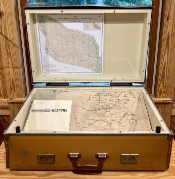 Color image of a brown suitcase on display at Prince William Forest Park's Visitor Center. The objects inside include the OSS Weapons Manual (1944) and two silk maps of China's rivers and railways developed by the recruits at the Chopawamsic training site. [Photo Credit: Dominique Mickiewicz]