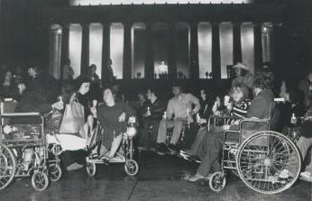 A black-and-white photo. A large group of people gathered in front of the Lincoln Memorial, it is dark outside. Most of the people in the foreground are sitting in wheelchairs and holding candles.