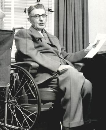 A black-and-white photo. Hugo Deffner, a white man in his 60s, sits in his wheelchair at a desk. He is holding a piece of paper in one hand and wearing a suit. He is looking at the camera.