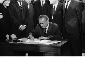 President Lyndon B. Johnson, leaning over a desk as he signs the 1968 Civil Rights Bill. Several men in suits surround him, watching.