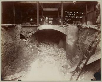 A black-and-white photo of the interior of Fords Theatre in 1893 looking from the basement up to the first floor. There is a pile of debris in the basement. There is a small group of people standing on the first floor. Handwritten on the photo is "view looking east from 10th street."