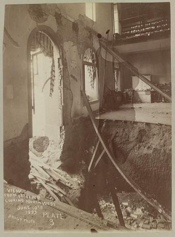 A black-and-white photograph of the interior of Fords Theatre after the collapse. A large window is blown out and the first and second floors are caved in. There is debris everywhere, paint peeling from the walls, and the curtains on the windows are tattered.