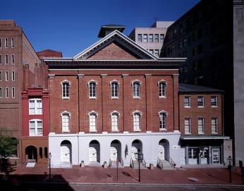 A color photo of the exterior of Fords Theatre and surrounding buildings ca. 1980-2006. The building is three stories, red brick, with tall windows across every floor. The roof is pointed.