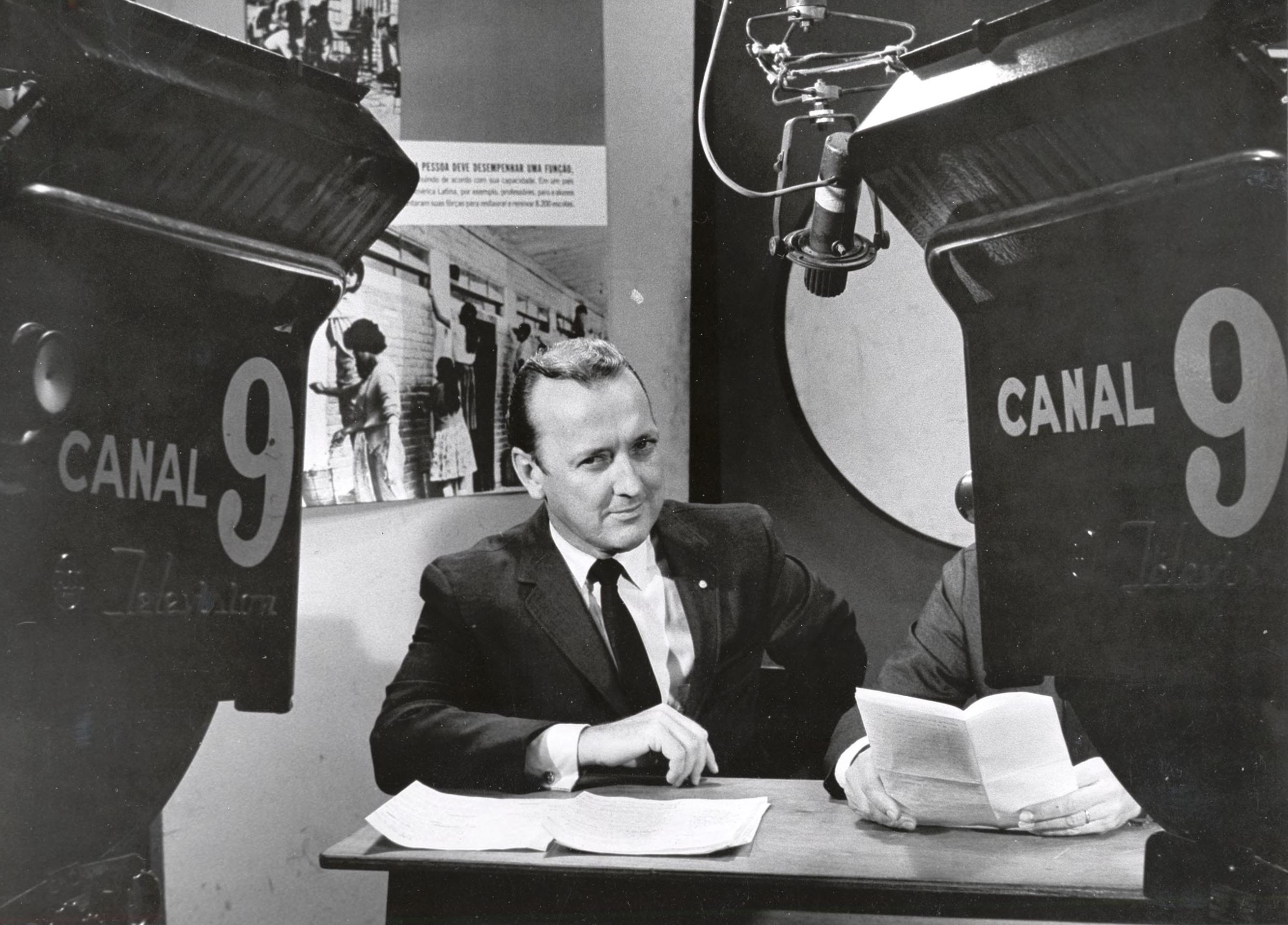 Grant sits at a desk in a newsroom. He is sharply dressed with a suit and tie. He looks intently at someone or something not captured in the picture. 