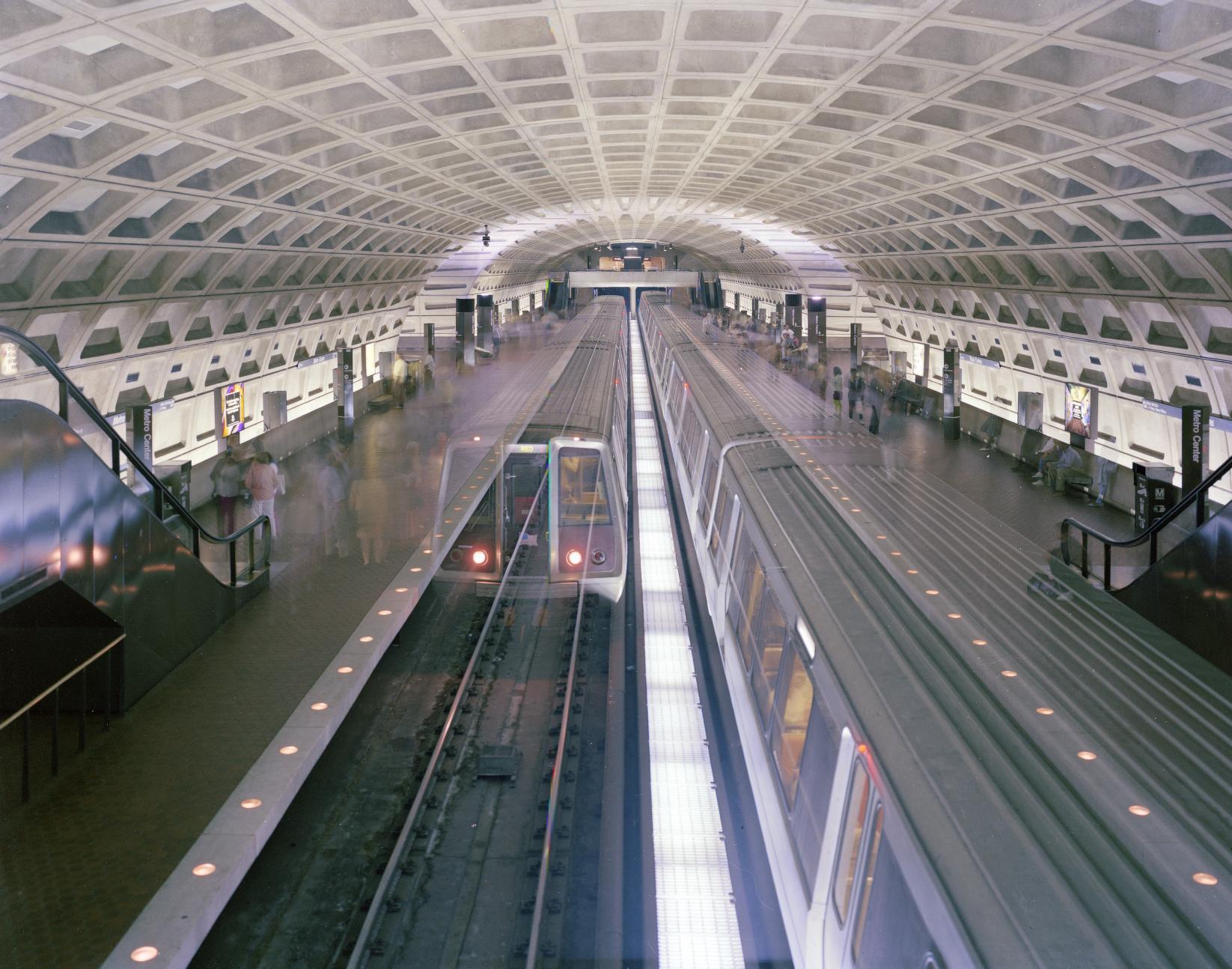 An overhead view of a DC Metro stop with trains running in either direction and blurry people moving around on the platforms. The signature egg carton-esque ceiling arches overhead.