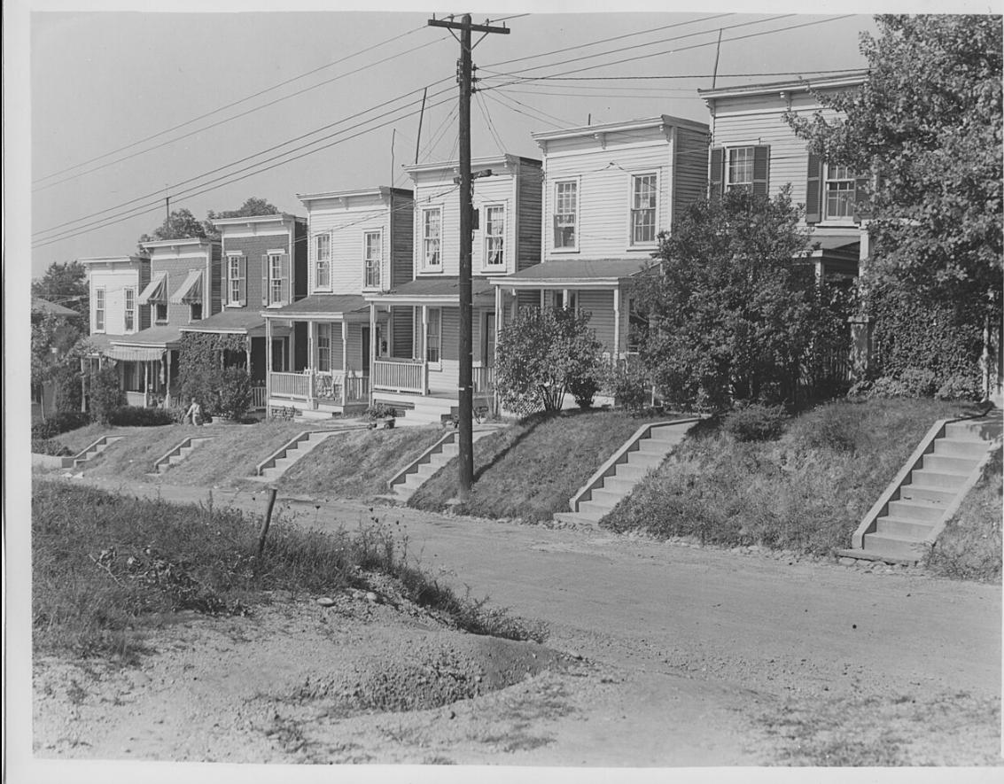 A black-and-white image of a street with 7 row houses on a hill. (Source: National Park Service Museum Resource Center, Public domain, via Wikimedia Commons) 