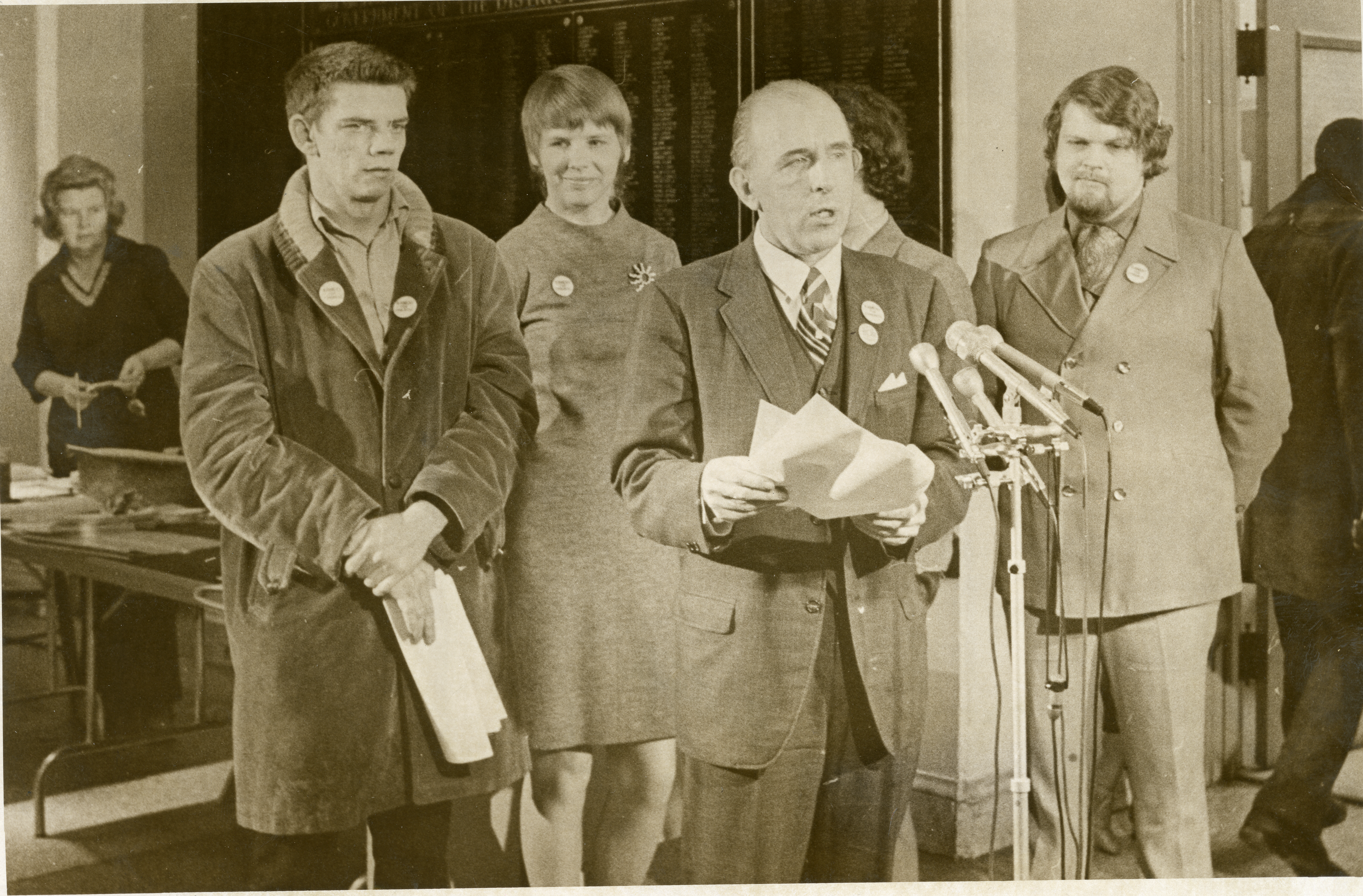 Frank Kameny holds papers and speaks into a group of microphones, flanked by two men and two women.