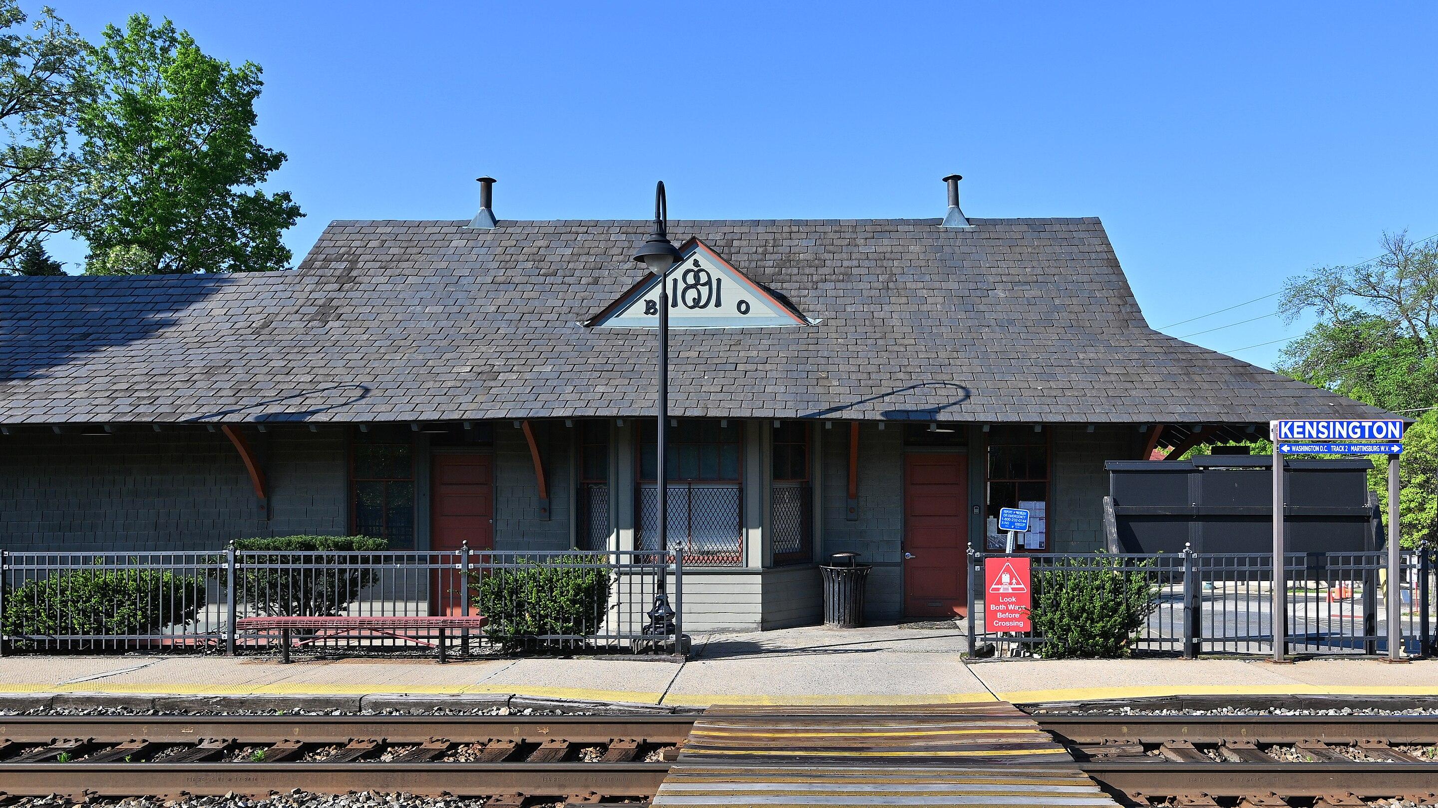 Kensington MARC Station in Maryland, showing a red brick station building with a steeply pitched roof and a prominent tower, alongside train tracks under a partly cloudy sky. A platform with a small shelter is visible, with trees and a few buildings in the background.(Source: G. Edward Johnson on Wikipedia. Cropped and used via Creative Commons Attribution 4.0 International License.)