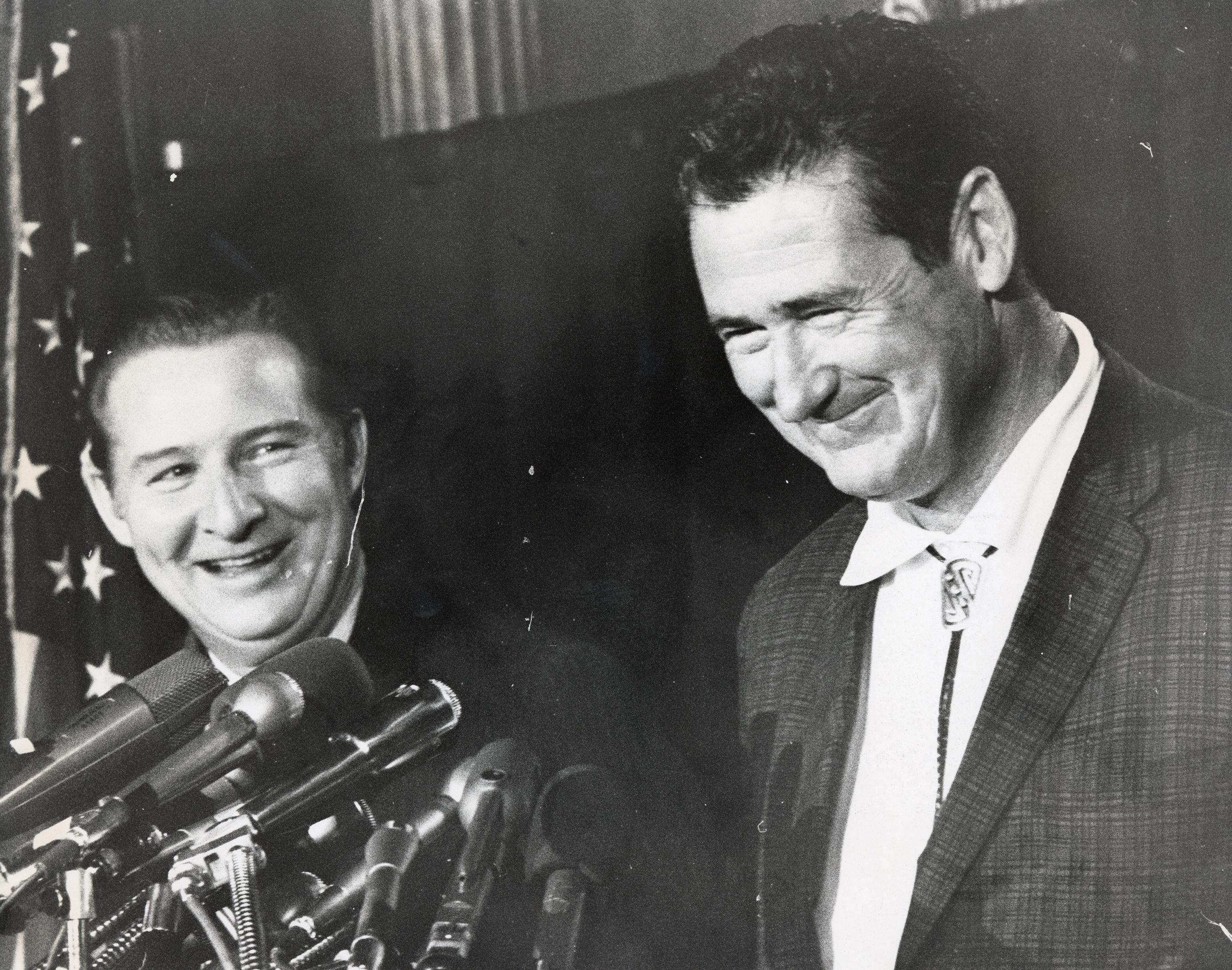 Bob Short and Ted Williams smile as they stand next to each other behind a collection of microphones.