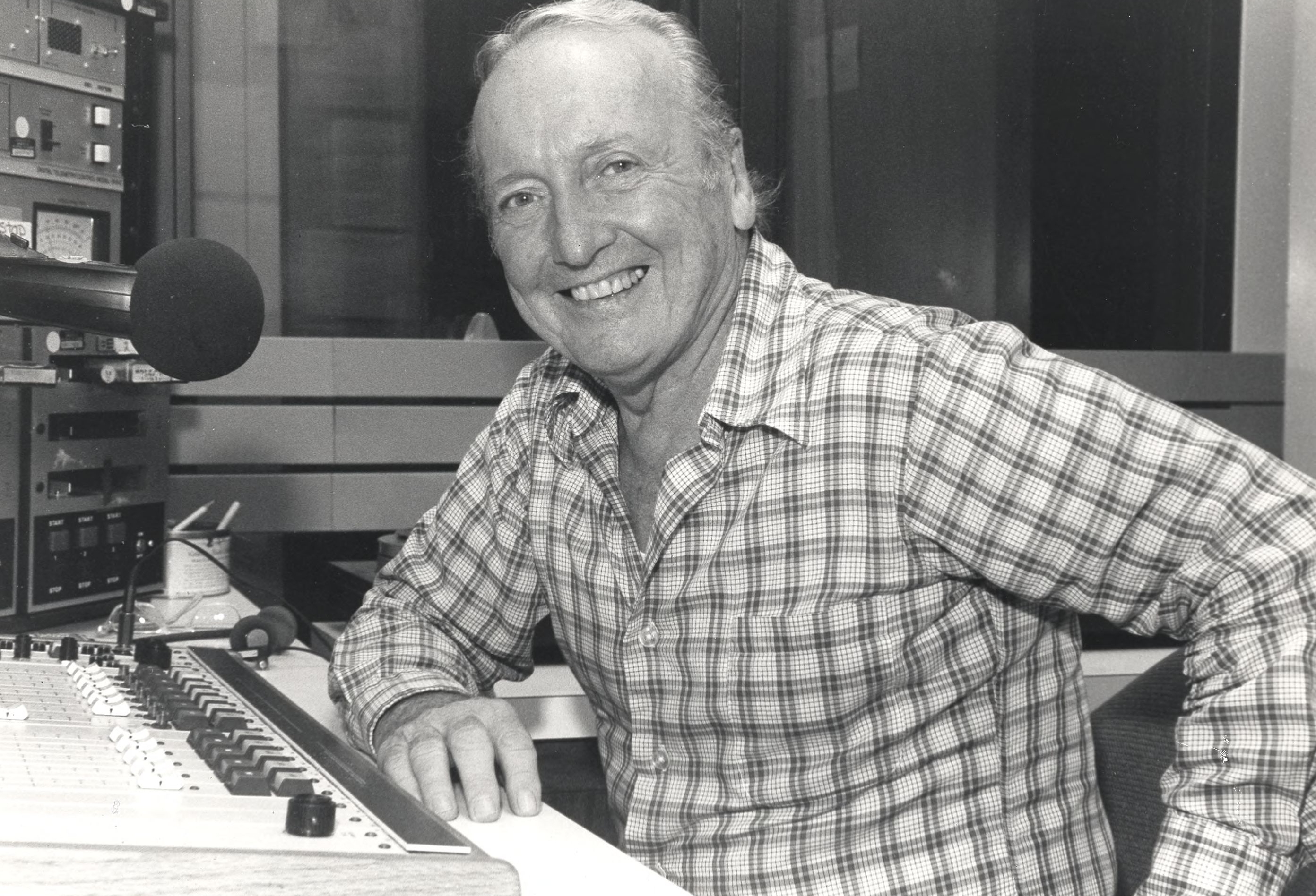An older Grant sits at a desk in a radio station booth. He has an elbow on the table and gives a big smile to the camera.